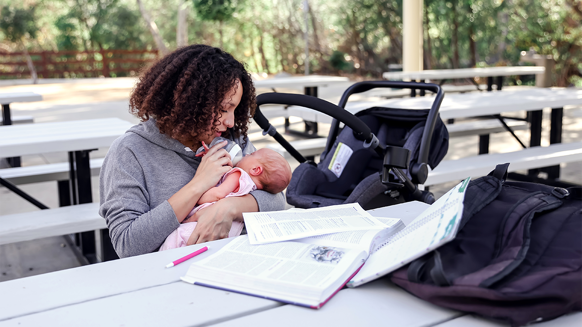 A young mom studies for a college exam outside at a picnic table while feeding her baby a bottle