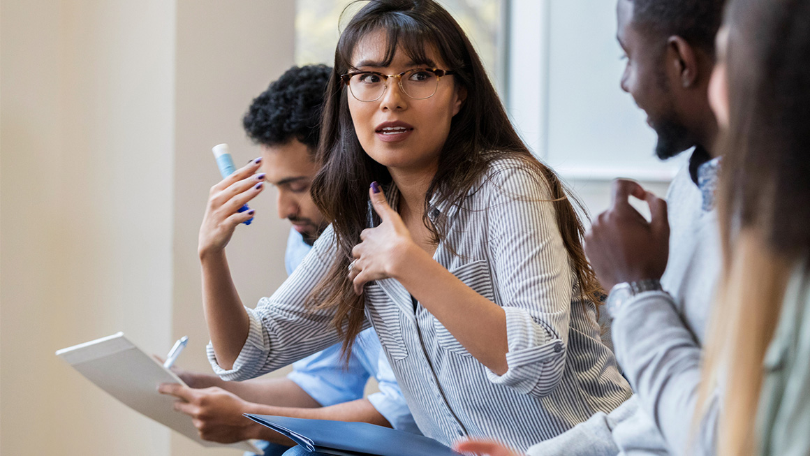 A woman with dark hair and glasses engages in a group discussion.