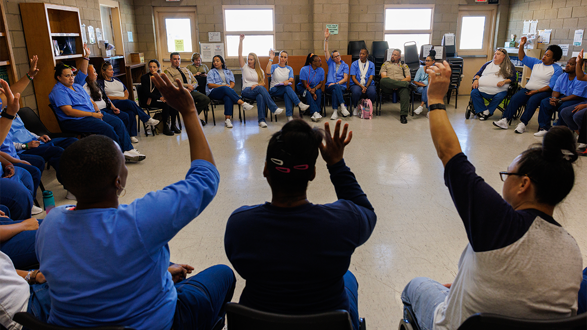 About two dozen incarcerated women sit in chairs arranged in a circle in a classroom. Some raise their hands.  