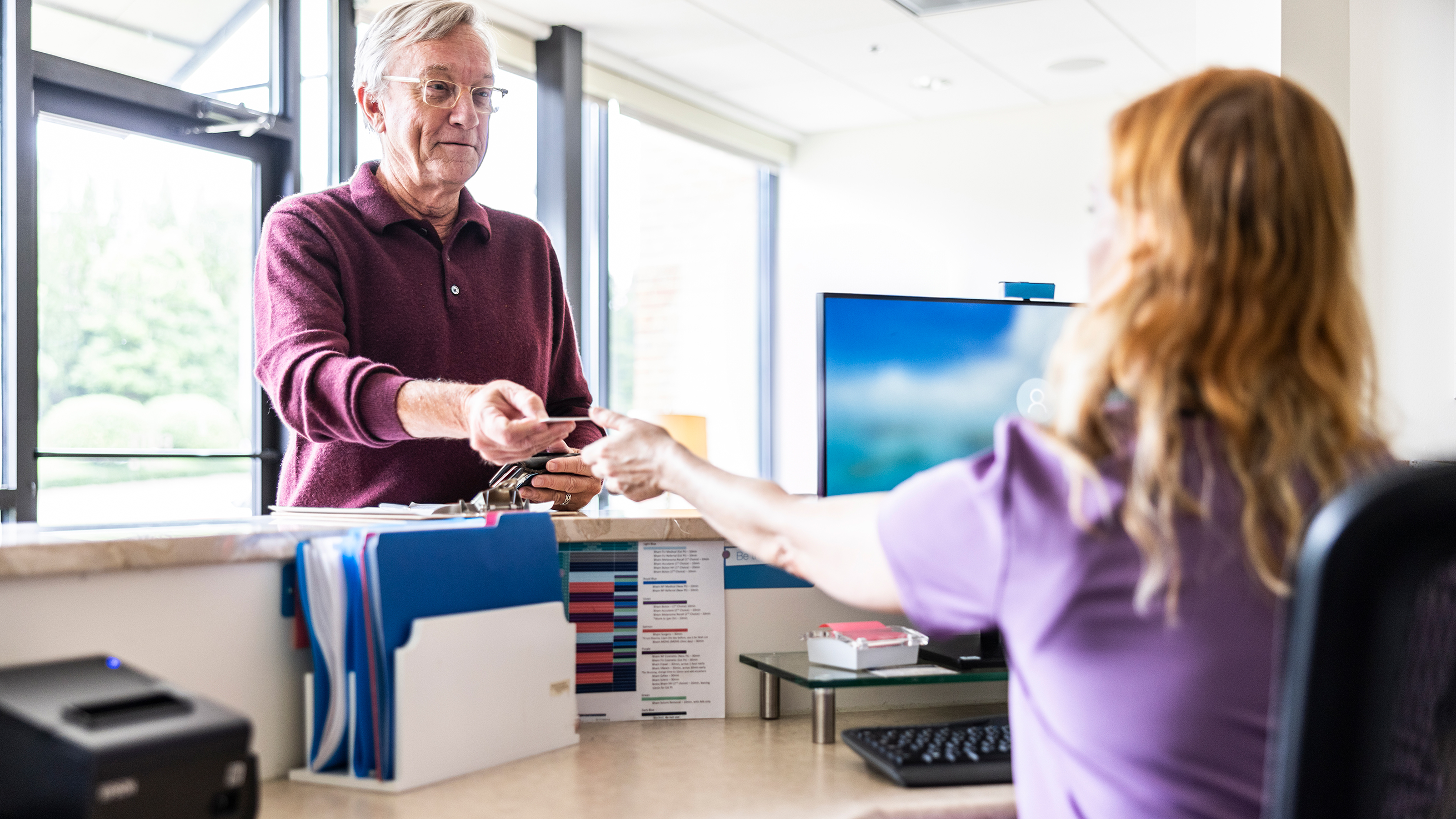 A man reaches forward and hands a card to a receptionist at a doctor's office
