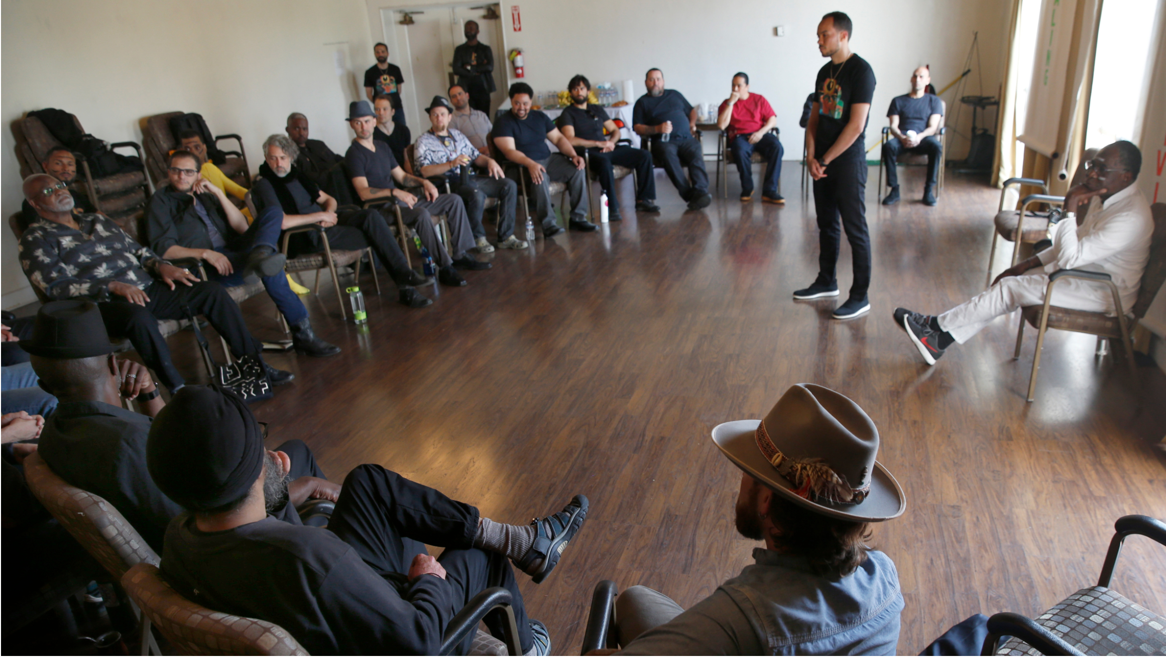 A group of men sits in chairs in a circle around one man in the middle