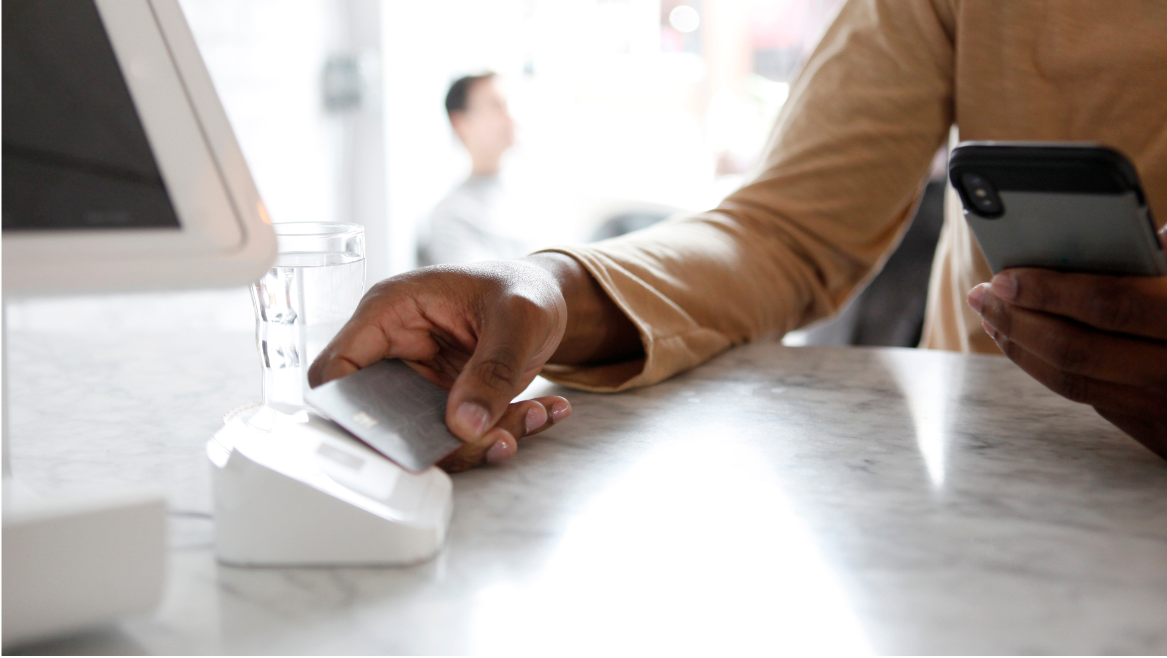 Photo of someone tapping their credit card on a credit card reader as they look at their phone. 