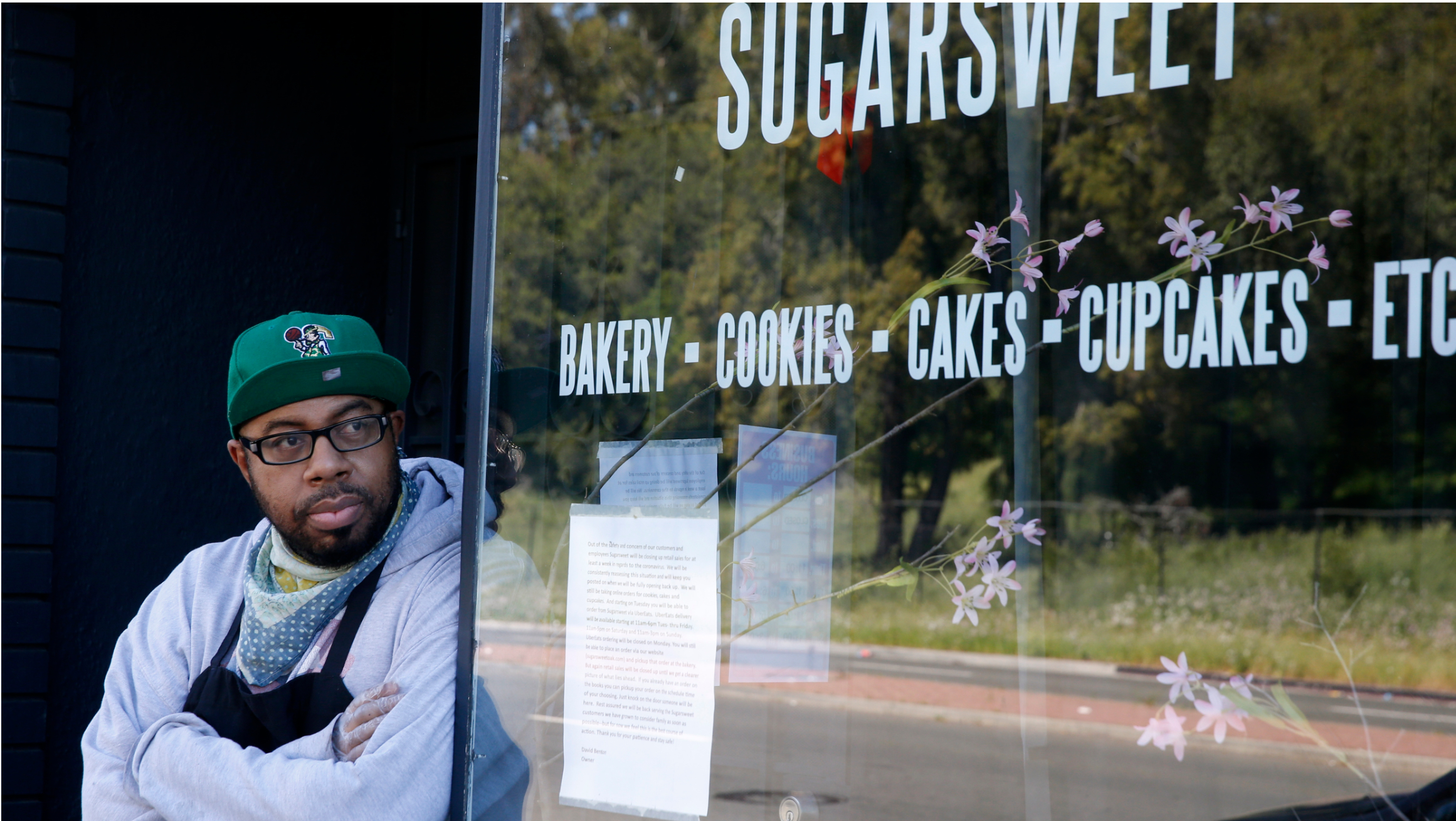 A man and small business owner leans against the doorway of a bakery looking off into the distance