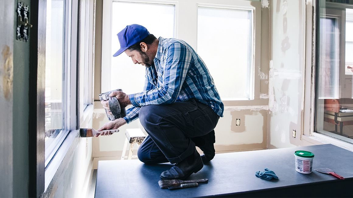Photo of a man kneeling on a table and painting window trim.  