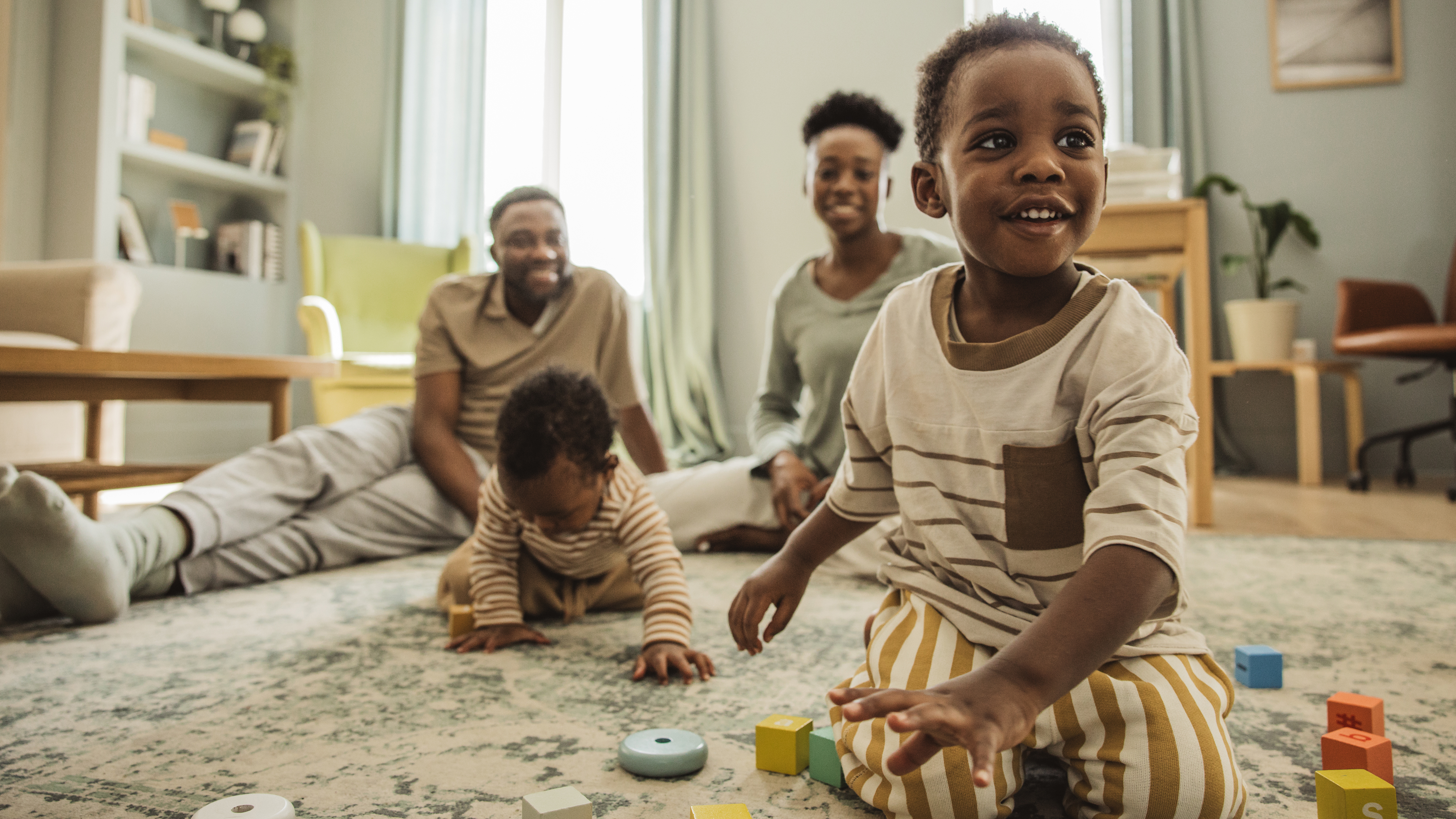 A family of two Black parents and two toddlers sits in their living room on the floor. The toddlers are playing with blocks.