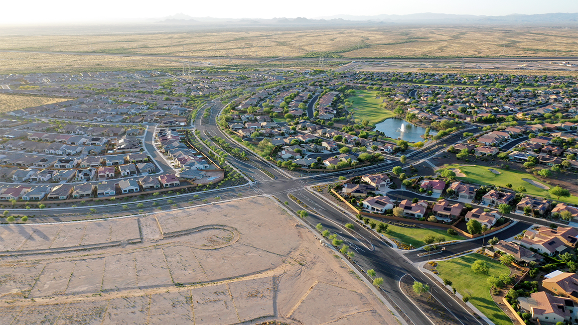 Aerial view of a large suburban neighborhood in a desert landscape, with curving residential streets, single-family homes, a small lake with a fountain, and a golf course, bordered by open undeveloped land and distant mountains.