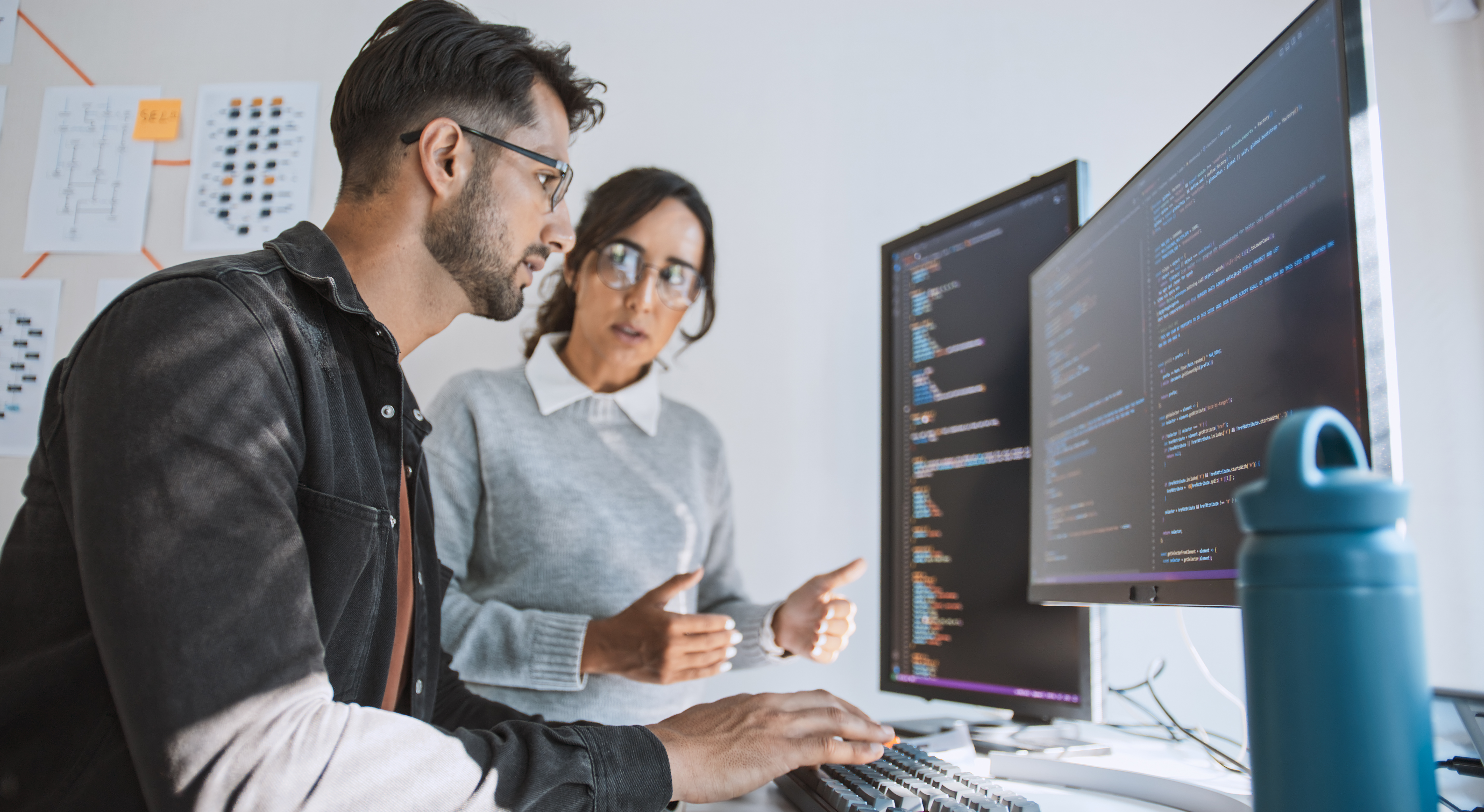 Photo of two young professionals looking at lines of code on two monitors.