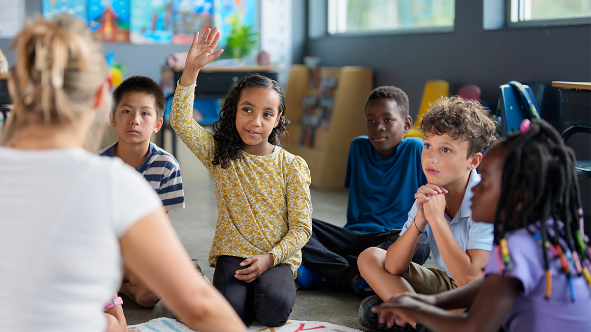 Photo of a young girl of color raising her hand as she and other students sit on a rug with their teacher. 