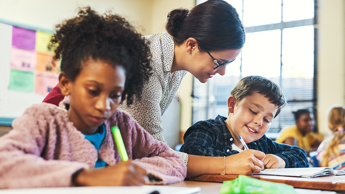    A teacher leans over a desk to help a student writing in a notebook, while another student beside them works independently.