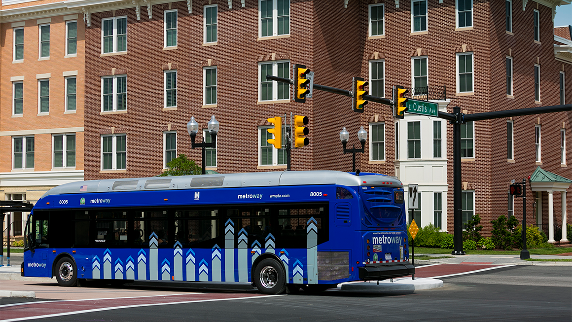 Blue Metroway city bus stopped at an intersection with traffic lights in front of a red brick building.