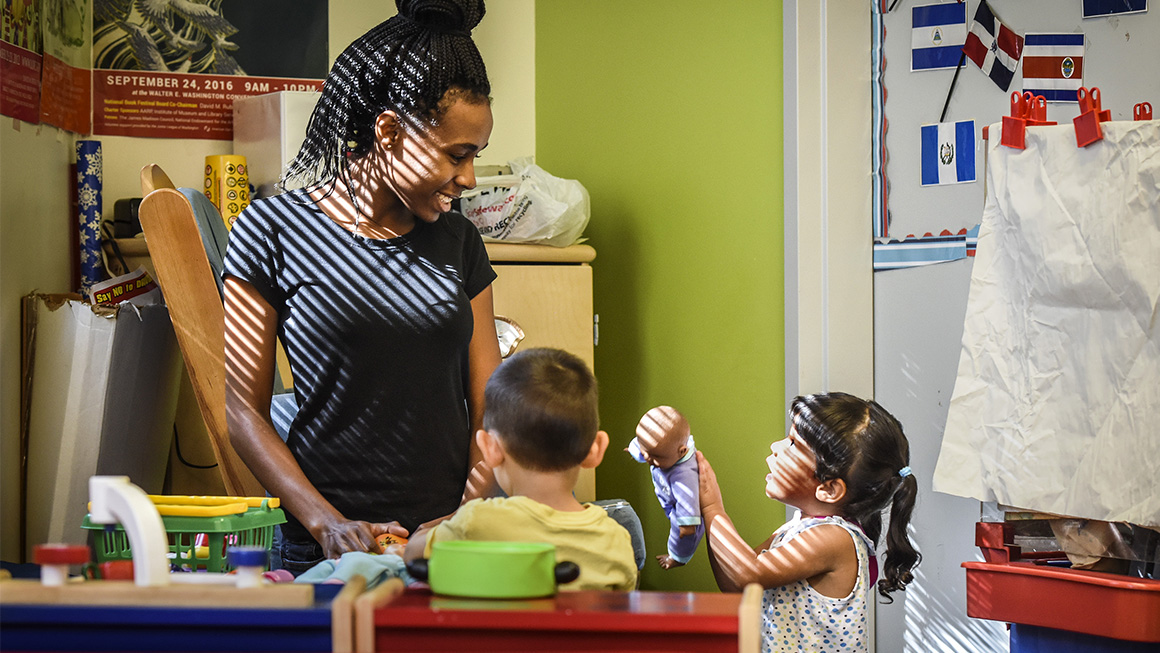 A child care provider smiles at two toddlers in a sunny classroom