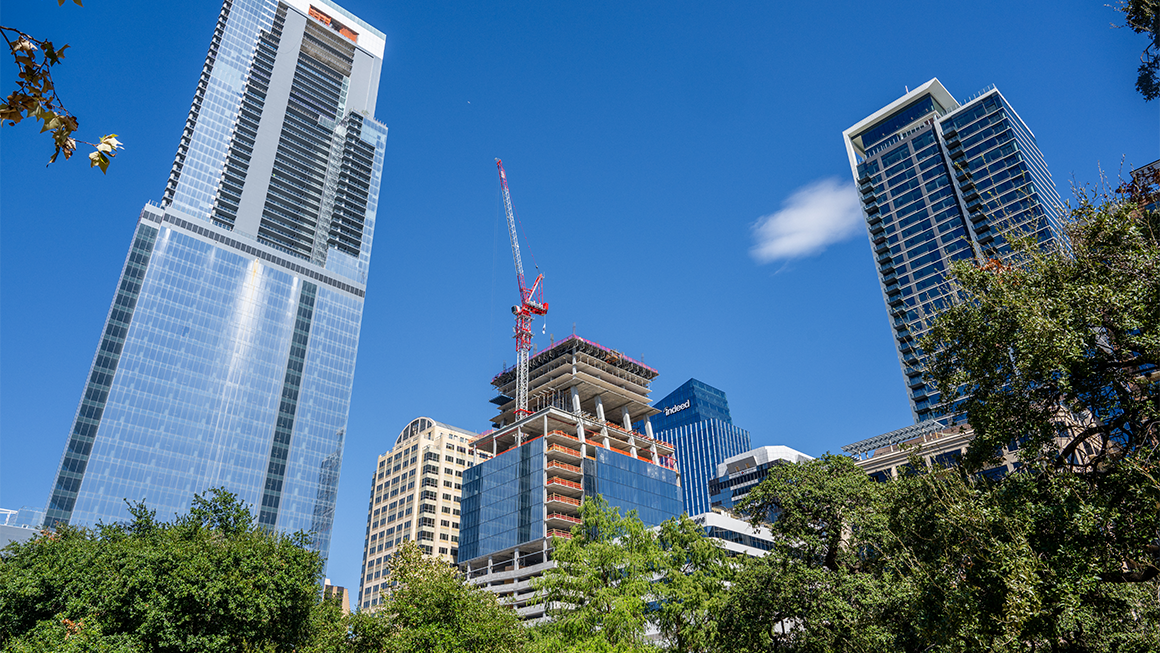City skyline with modern glass skyscrapers and a high-rise under construction with a crane, surrounded by trees under a clear blue sky.