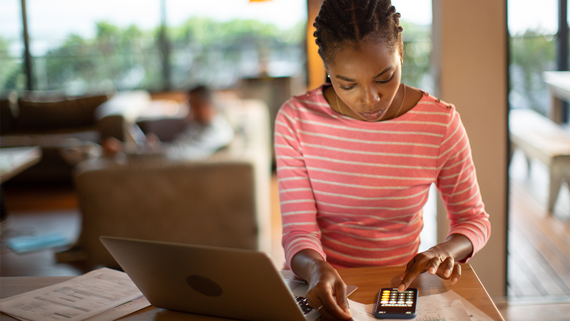 Photo of a young Black woman sitting in front of her laptop and using the calculator on her phone.