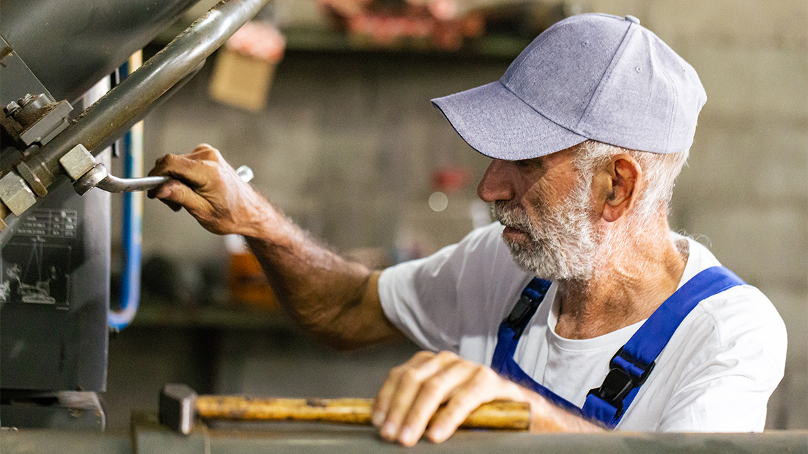 Person in a workshop operating industrial machinery while wearing a cap, white shirt, and blue overalls.