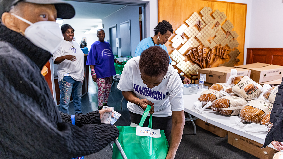 People collect food items at a community distribution table, with volunteers placing packaged items into a green bag; boxes and loaves of bread are visible nearby.