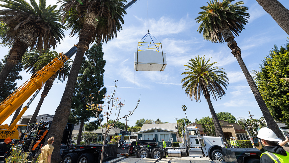 Crane lifting a large prefabricated structure over a residential street lined with tall palm trees and work trucks.