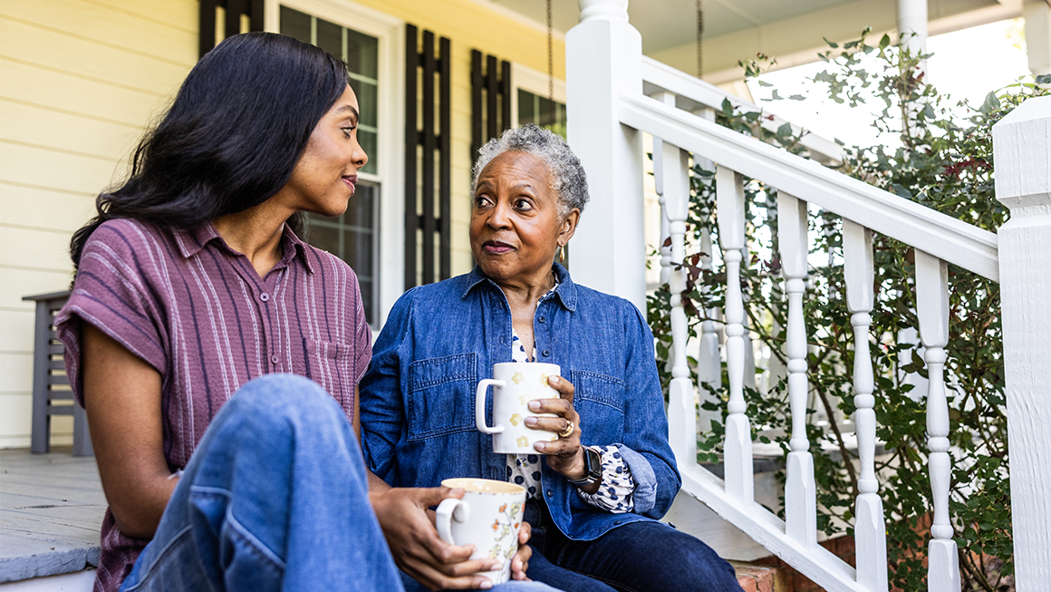Image of an older woman of color and her daughter sitting on porch steps and drinking a warm beverage.  