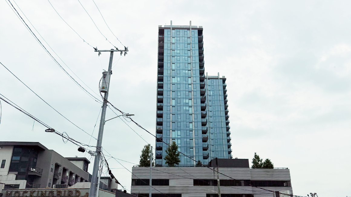 Modern blue-glass high-rise apartment building with balconies, viewed from street level alongside power lines and a utility pole under a cloudy sky.