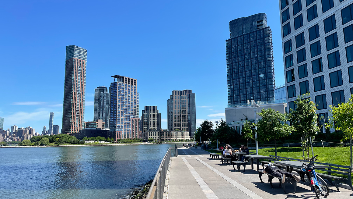 Waterfront walkway with benches and people, bordered by modern high-rise buildings along a river under a clear blue sky.