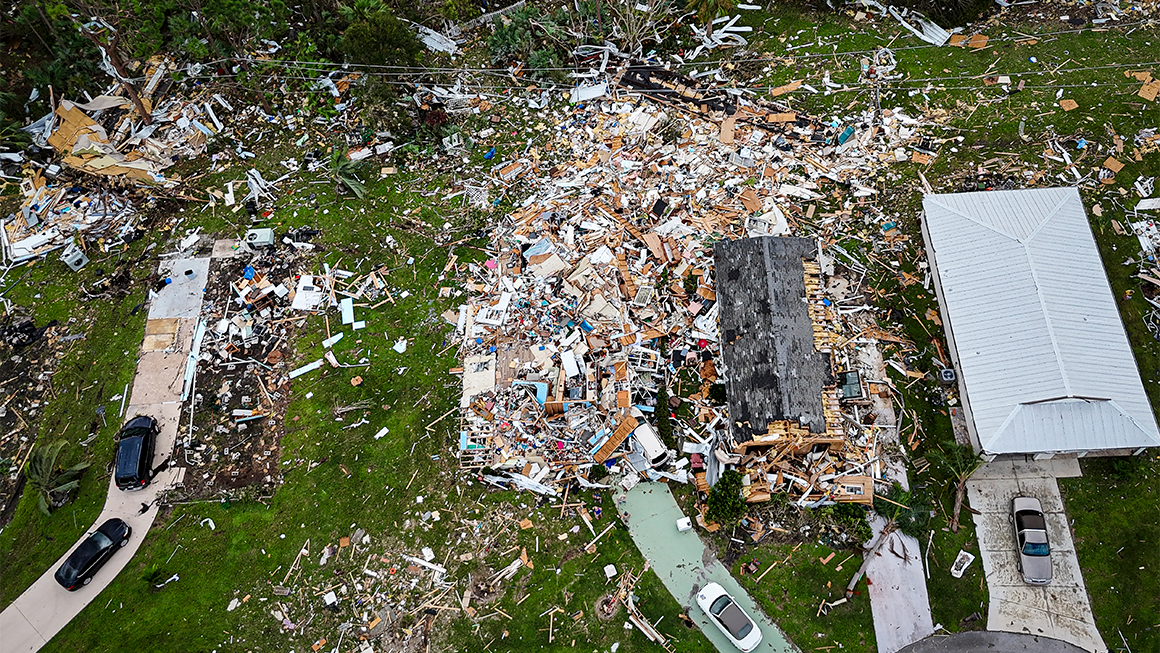 Aerial view of a neighborhood showing severe storm damage, with one home reduced to debris and wreckage scattered across surrounding yards.