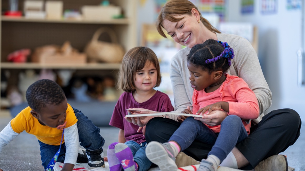 An adult sits with three young children on a classroom floor, reading a book together during a group learning activity.
