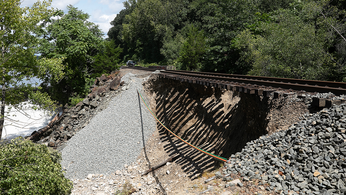 A section of railroad track where the supporting embankment has washed away, leaving the rails and wooden ties suspended above an eroded slope.