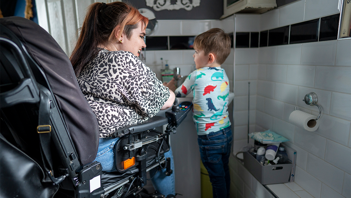 Image of a young disabled woman in a wheelchair helping her son wash his hands in the bathroom sink.