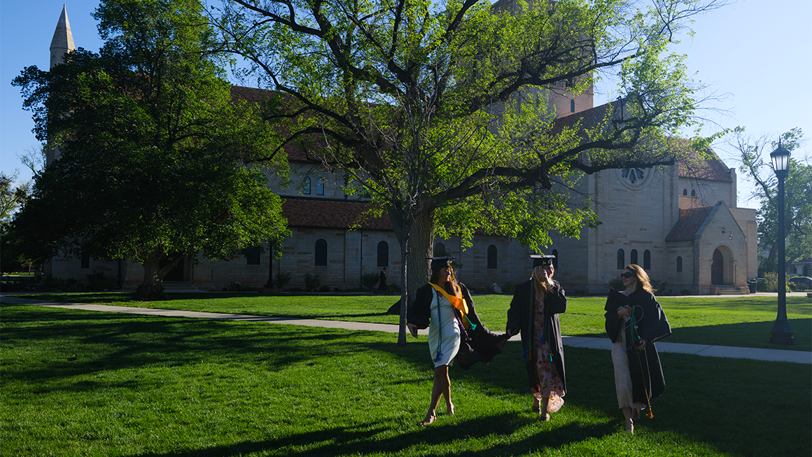 Three graduates in caps and gowns walk across a sunlit lawn in front of a large stone campus building and leafy trees.
