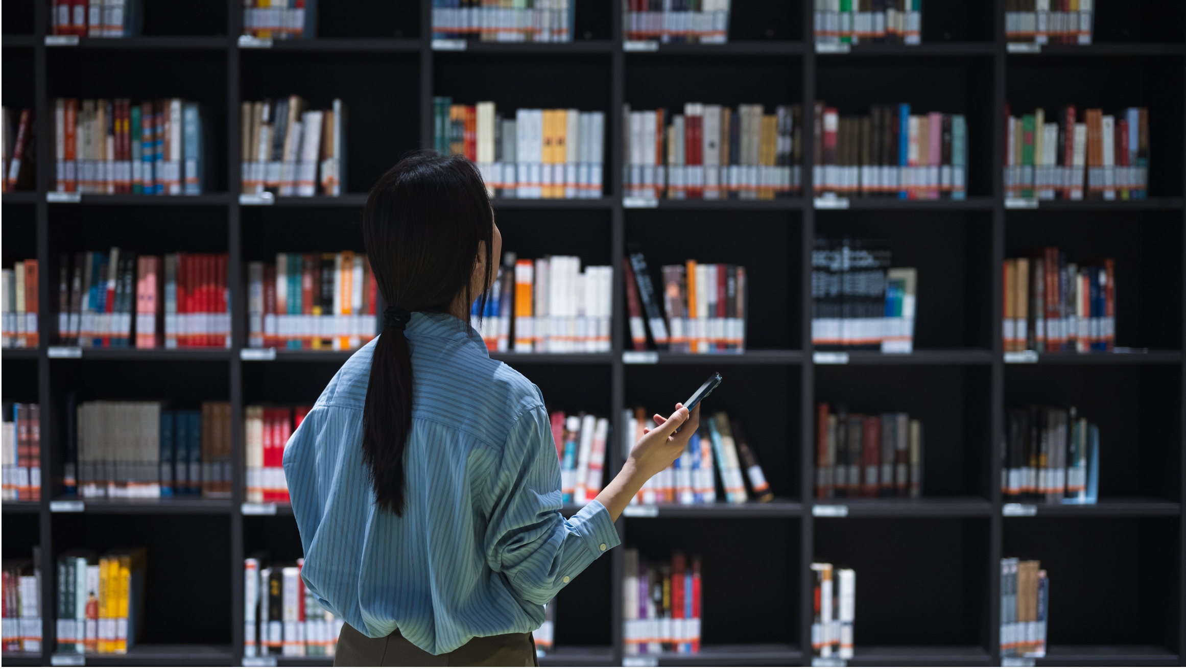 Person standing in front of tall library bookshelves, holding a smartphone while browsing rows of books.