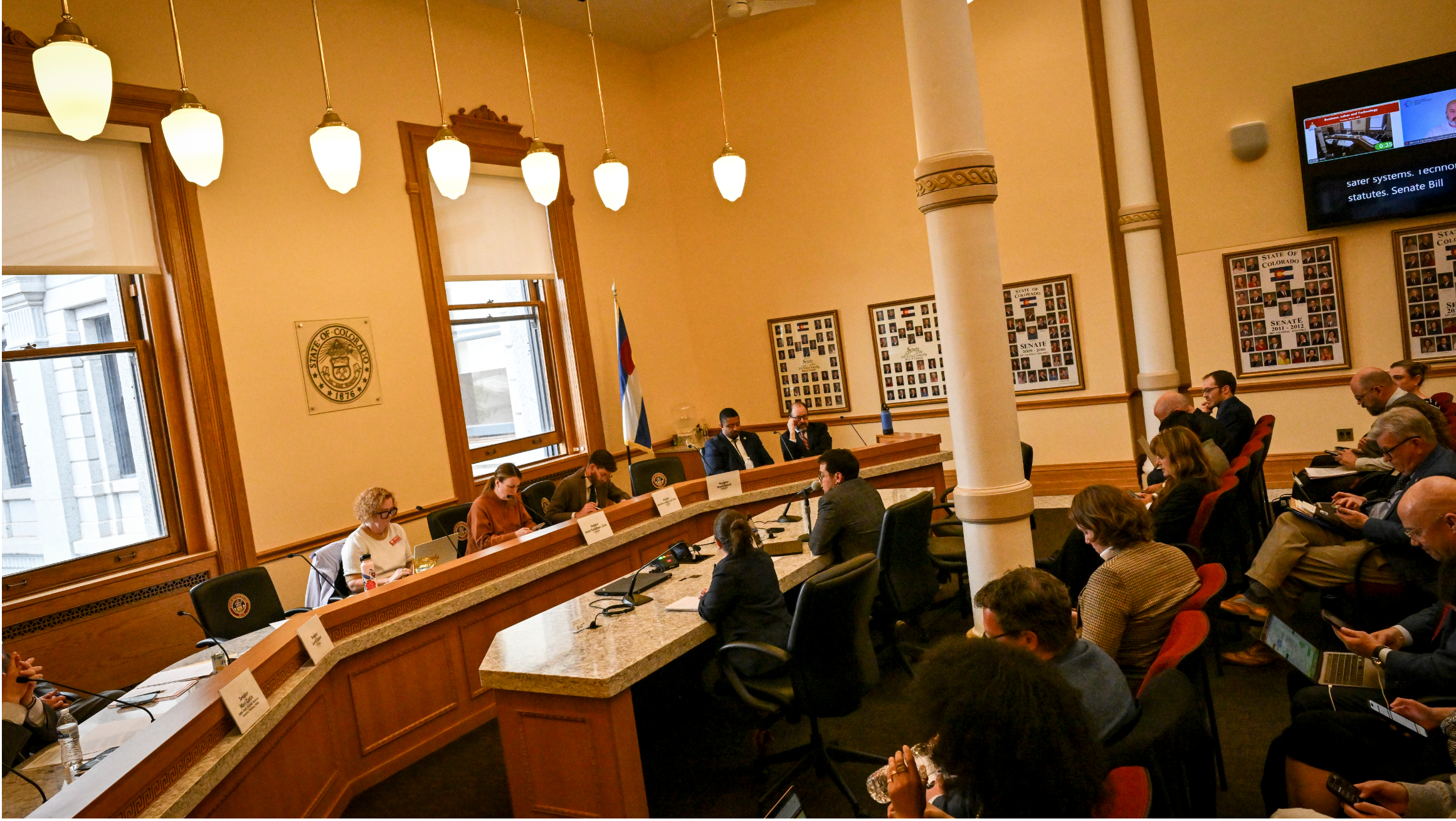 People seated around a curved committee dais during a legislative hearing in a large government meeting room with high ceilings, tall windows, and a Colorado state flag.