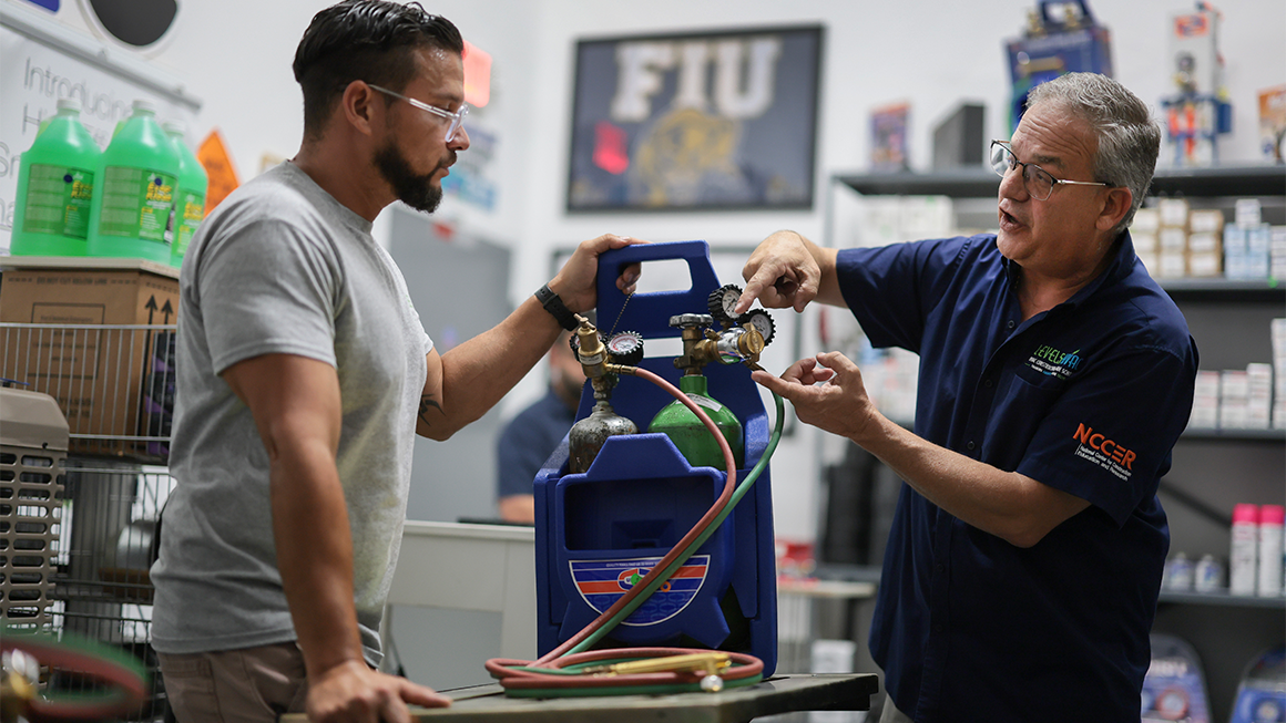 Two people in a workshop review HVAC refrigerant gauges connected to gas cylinders during hands-on technical training.