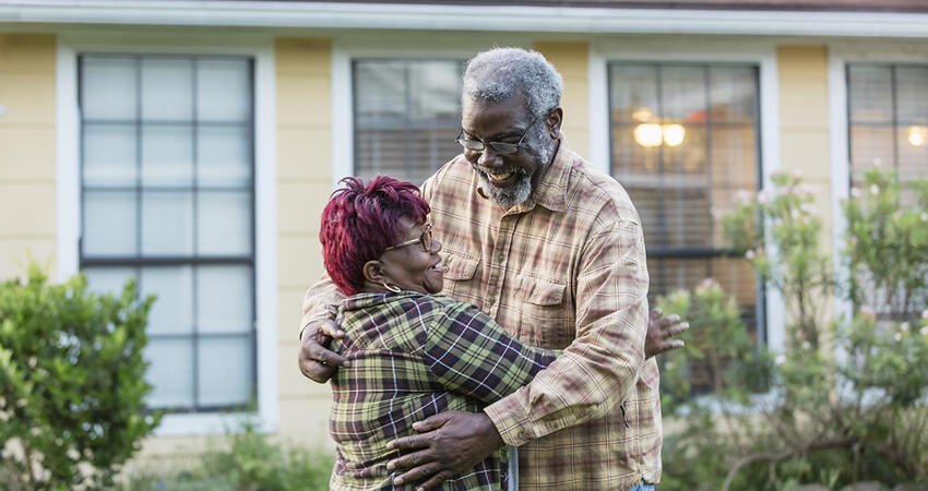 An older black couple hugging in front of housing.