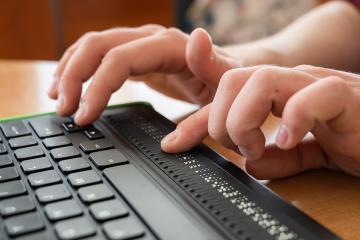 A blind man uses a computer with a Braille display and a computer keyboard. Inclusive device.