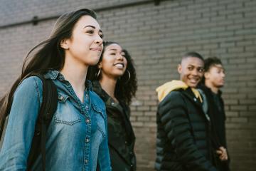 A smiling group of young adults walk to campus for classes on a sunny day, having fun as they enjoy the weather. Mixed ethnic group, wearing backpacks and shoulder bags with their text books.