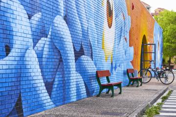 A colorful blue and orange mural covers the brick wall running alongside a street, with a couple benches and a bike rack.