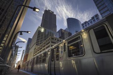 A close upward angle of a silver public train against a backdrop of skyscrapers and a whispy sky.