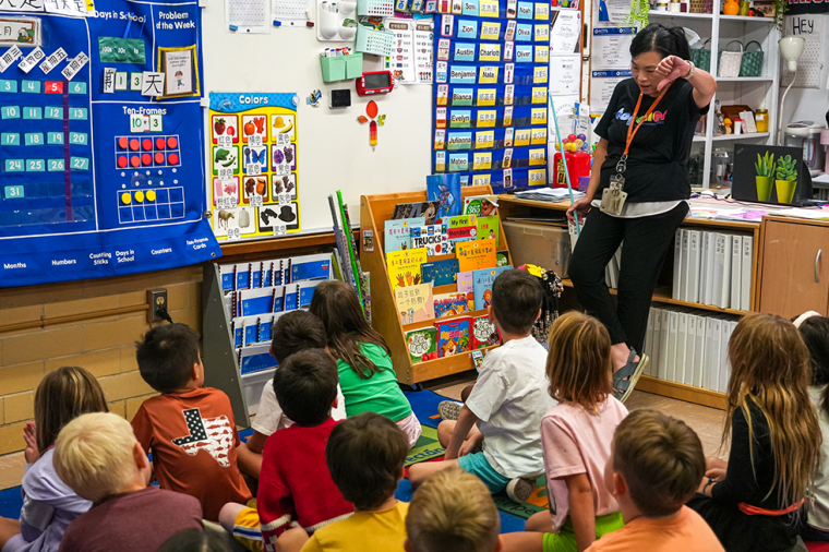 An educator stands at the front of a colorful classroom, reading to a group of young children seated on a carpet, surrounded by learning charts, books, and classroom displays.