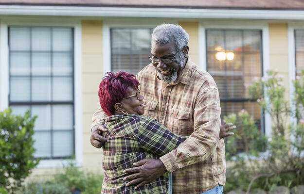 An older black couple hugging in front of housing.