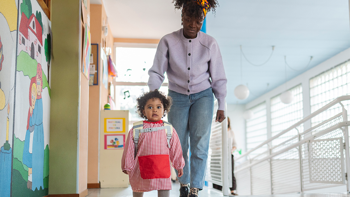 A young child walking through the halls of a day care, followed by her mother.