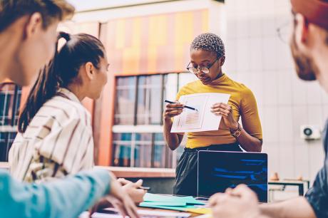 Coach holding paper sketch in hands and explaining data of diagram during workshop for students sitting at one table