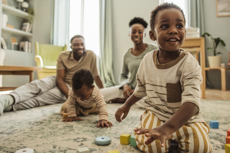 A family of two Black parents and two toddlers sits in their living room on the floor. The toddlers are playing with blocks.