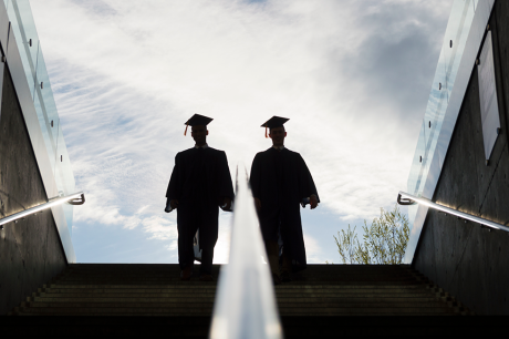 Two graduates in caps and gowns walk down a wide outdoor staircase, silhouetted against a bright sky, with glass railings and concrete walls on either side.