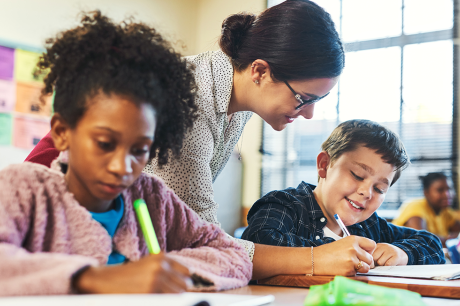    A teacher leans over a desk to help a student writing in a notebook, while another student beside them works independently.