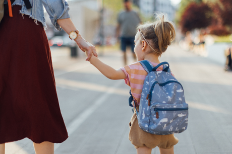A young child wearing a small blue backpack holds an adult’s hand while walking down a sunlit city sidewalk