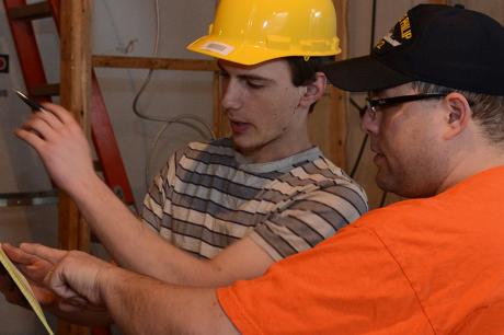 Two construction workers review a checklist inside a framed building, one pointing at a wall.