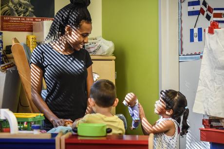 A child care provider smiles at two toddlers in a sunny classroom