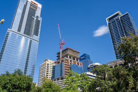 City skyline with modern glass skyscrapers and a high-rise under construction with a crane, surrounded by trees under a clear blue sky.