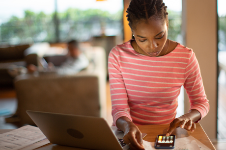 Photo of a young Black woman sitting in front of her laptop and using the calculator on her phone.
