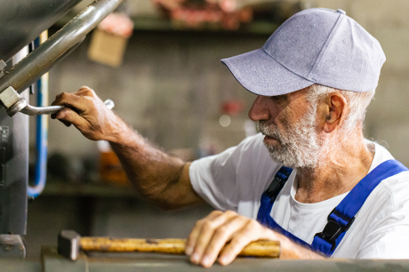 Person in a workshop operating industrial machinery while wearing a cap, white shirt, and blue overalls.
