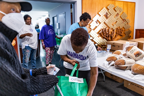 People collect food items at a community distribution table, with volunteers placing packaged items into a green bag; boxes and loaves of bread are visible nearby.